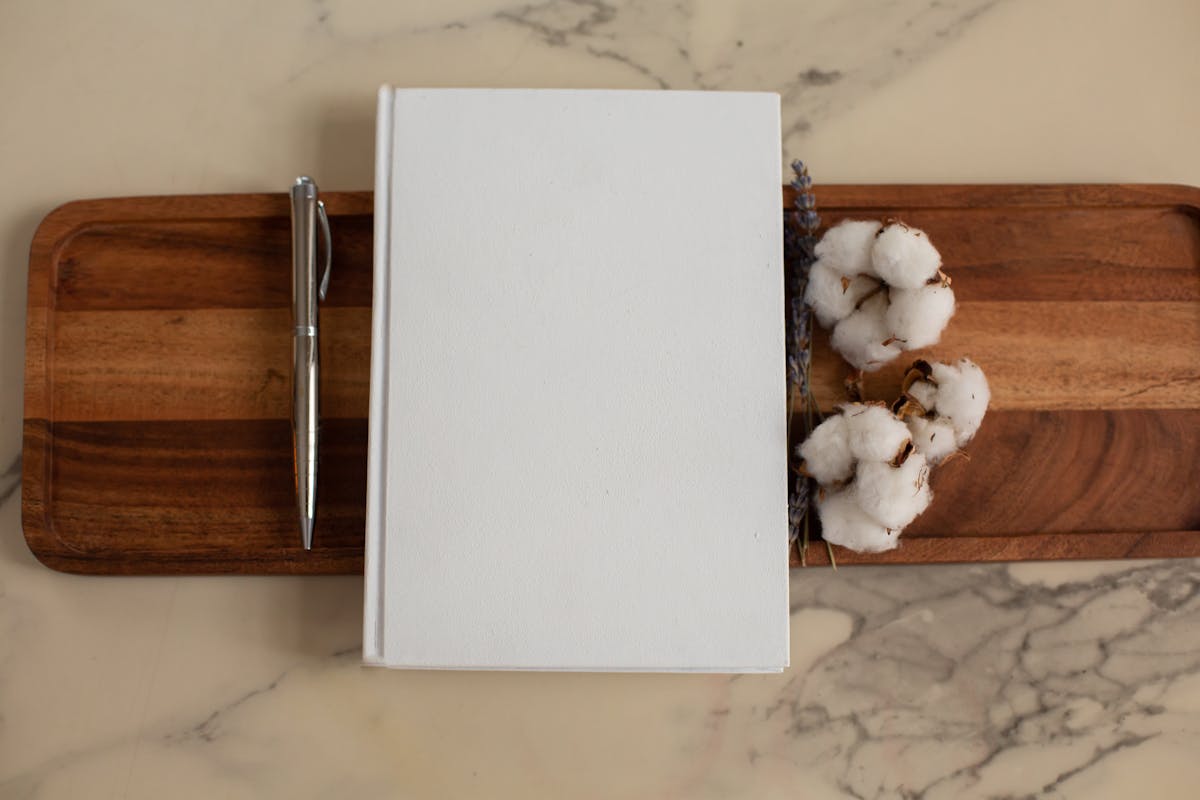 Overhead view of composition of empty hardcover book lying between metallic pen and soft cotton flowers on wooden tray with recesses