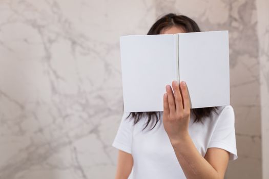 Unrecognizable female in casual t shirt with opened white mock up notebook in hands standing in light room near wall
