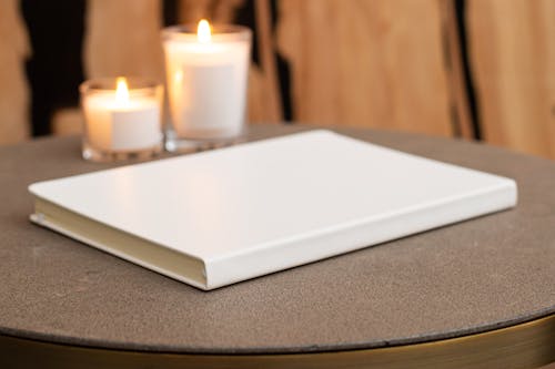 Blank stylish white notebook on round table with burning candles near wall in light room on blurred background at home