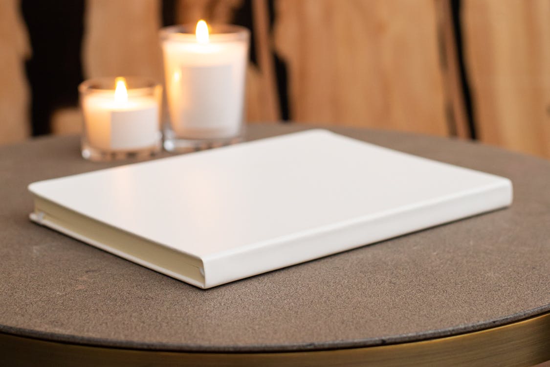 Blank stylish white notebook on round table with burning candles near wall in light room on blurred background at home