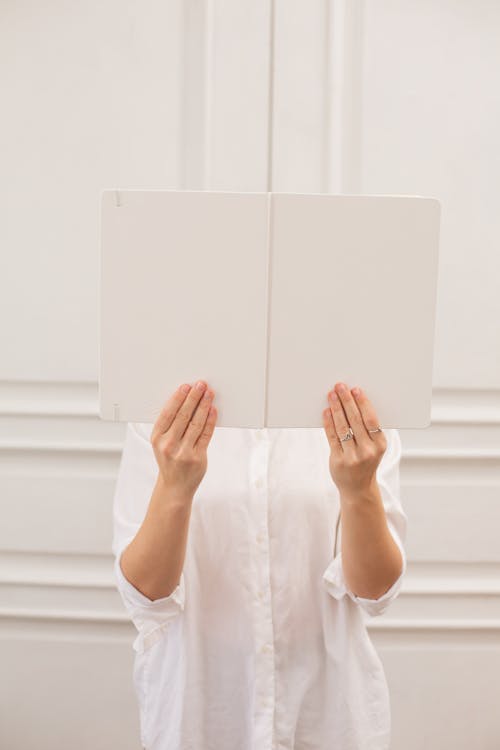 Free Unrecognizable female in white blouse hiding face with opened mock up notepad while standing in light room near door in apartment Stock Photo