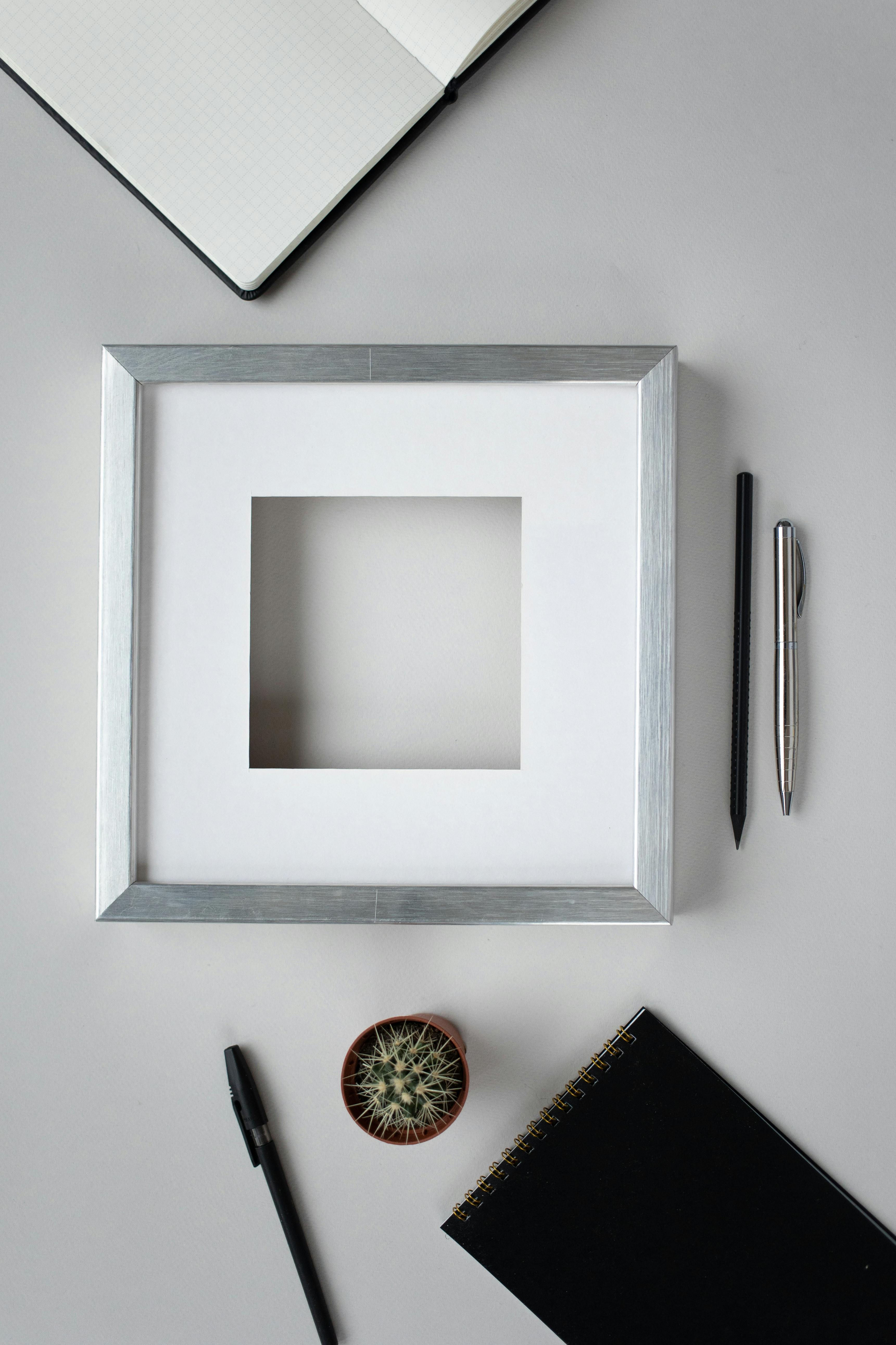Top view of mock up square shaped frame frame placed on table with pens and notebooks near cactus in light room
