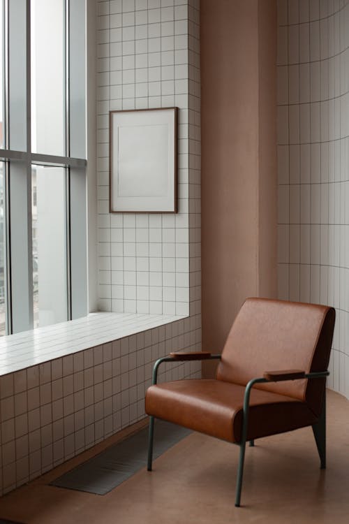 Free Blank frame hanging on tiled wall near window in small light room with soft brown leather armchair on floor at home Stock Photo