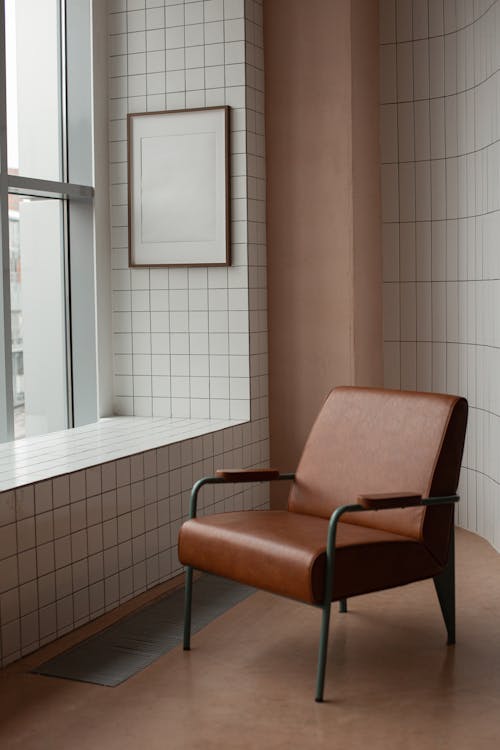 Comfortable brown armchair placed near tiled wall with mock up frame hanging near window in light small room at home