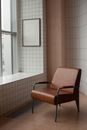 Comfortable brown armchair placed near tiled wall with mock up frame hanging near window in light small room at home