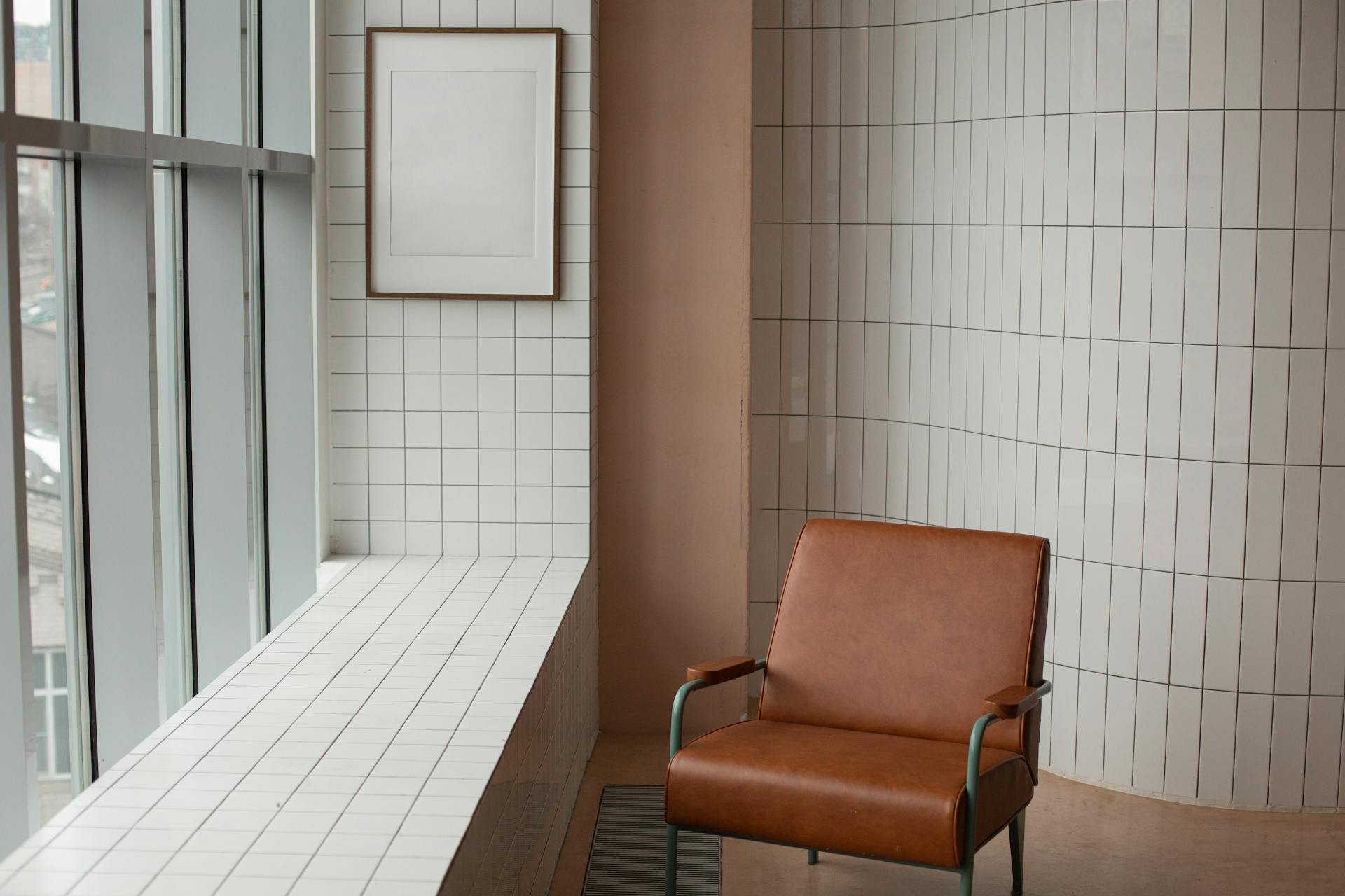 Brown armchair placed near mock up frame hanging near window and windowsill in small room with tiled walls at home