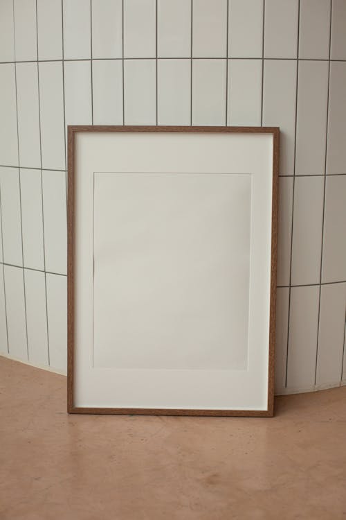 White rectangular blank frame with brown border placed on floor near wall with ceramic tiles in light room at home