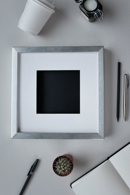 Top view arrangement of empty photo frame placed on white table near opened notebook pens and potted cactus