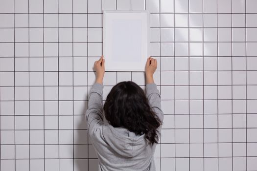 Back view anonymous brunette in gray hoodie hanging white empty photo frame on light tiled wall