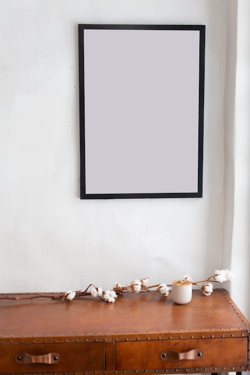 Empty black photo frame hanging above wooden cabinet with decorative twig in light bedroom