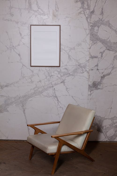 Empty white wooden photo frame hanging on marble wall near comfy armchair in light room