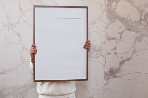 Crop anonymous person demonstrating white empty frame in hands near gray wall with patterns