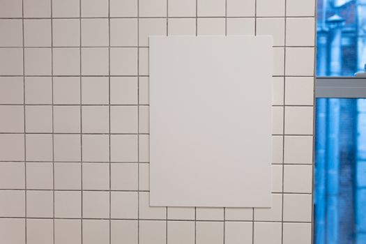 Blank white placard hanging on wall covered with white tiles inside of building