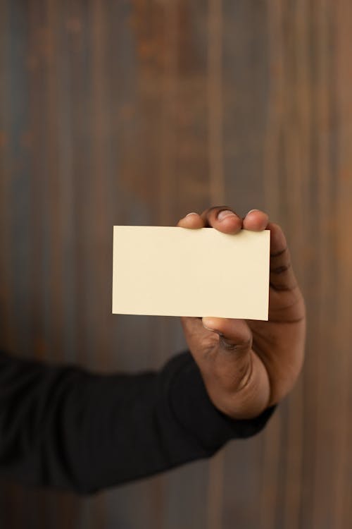 Unrecognizable African American male showing mock up business card with copy space in hand while standing near wooden wall in light room