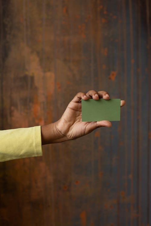 Anonymous African American person showing green blank business card with copy space in hand while standing near wooden wall in studio