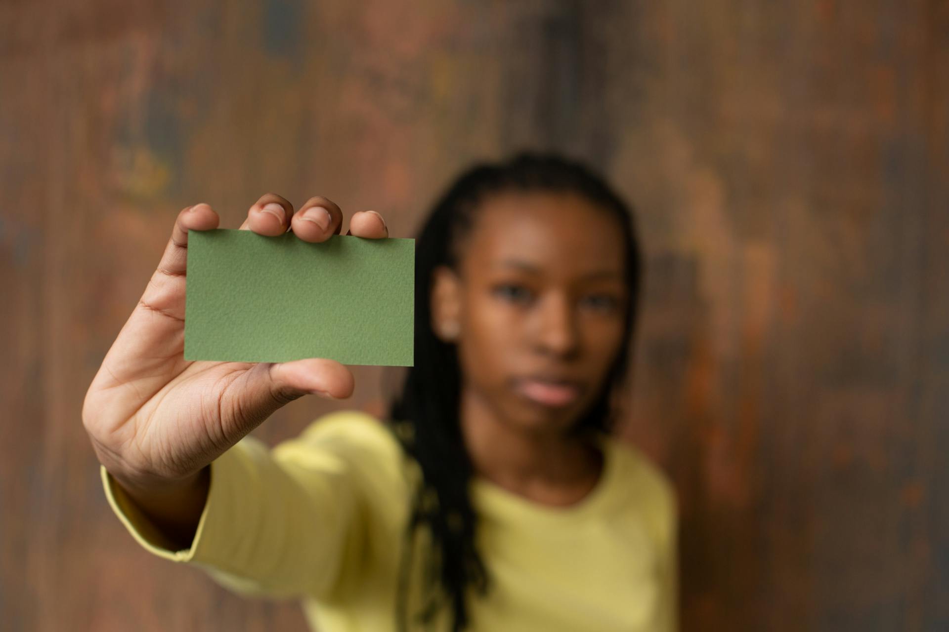 Soft focus of African American female looking at camera and demonstrating green mock up business card in hand while standing near wooden wall