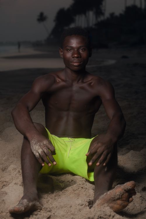 Free A shirtless black man sits on the sandy beach at twilight wearing bright swim shorts. Stock Photo