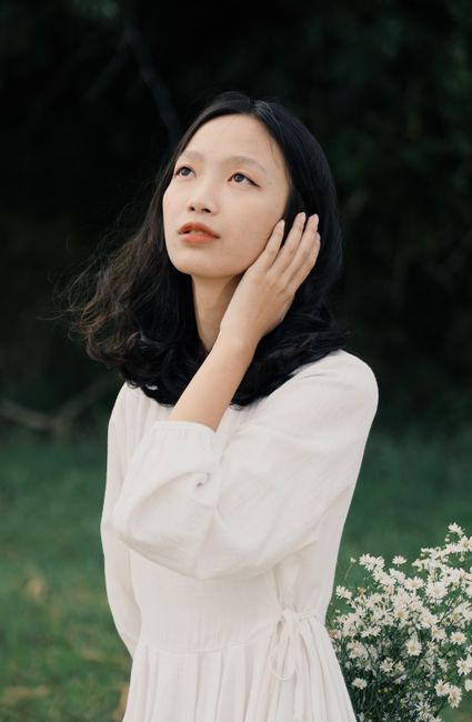 Dreamy Asian female in dress touching hair standing near blooming flowers and looking away
