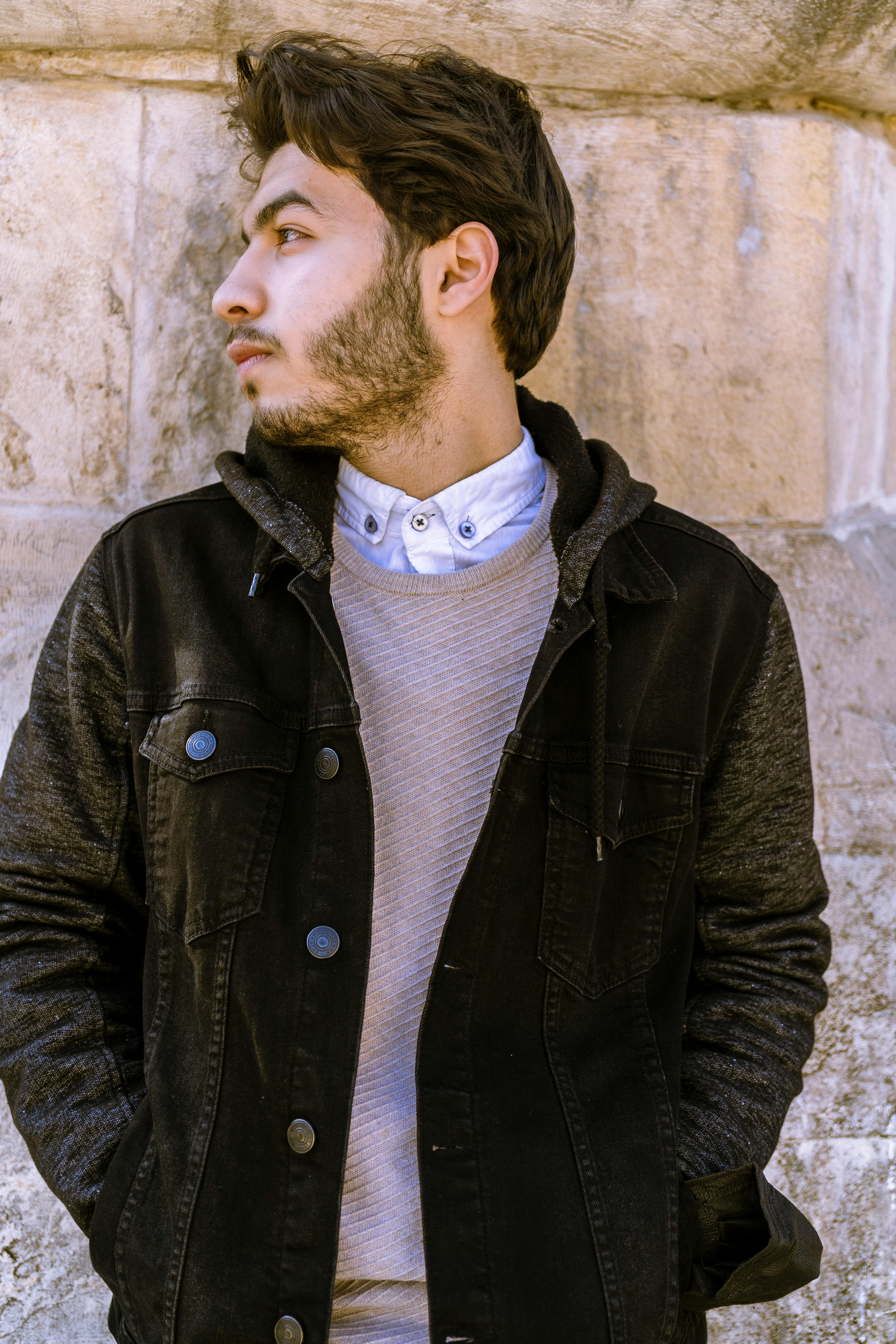 Stylish young man in a black jacket gazing thoughtfully against a stone wall background.