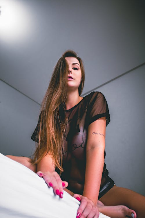 Low angle of self esteem young lady with long straight hair in translucent nightgown relaxing on bed and looking at camera