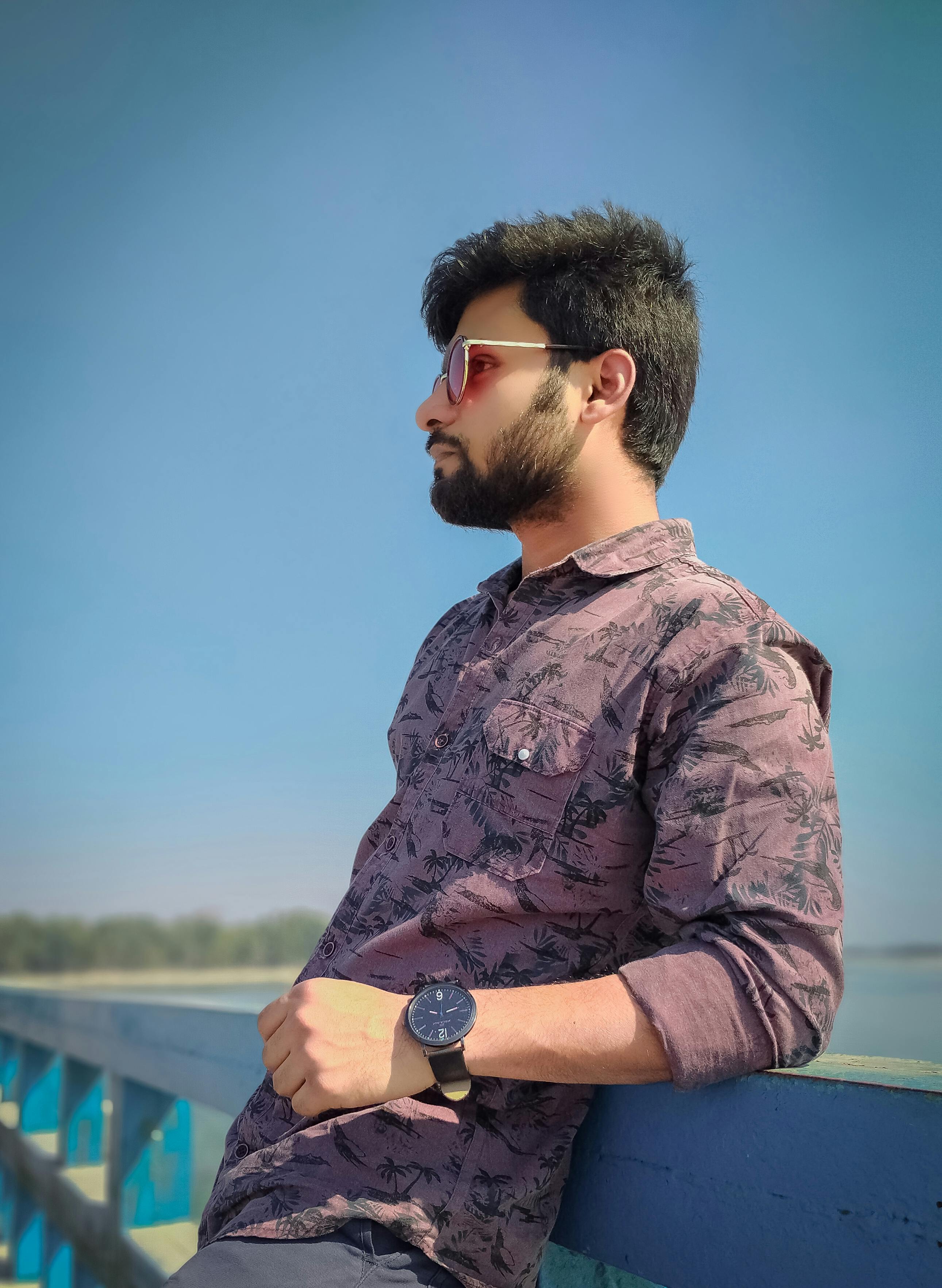 Young bearded man wearing sunglasses poses outdoors against a blue sky.