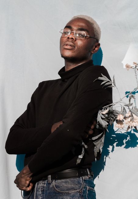 Self assured young African American male with dyed hair in trendy outfit and eyeglasses leaning on white wall with bunch of flowers in hand and looking at camera in sunlight