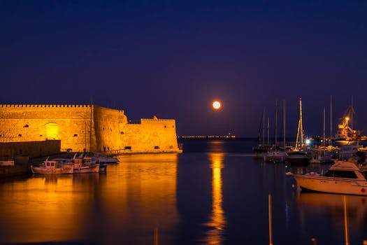 Moonlit harbor scene with boats and illuminated fortress reflecting on the water.