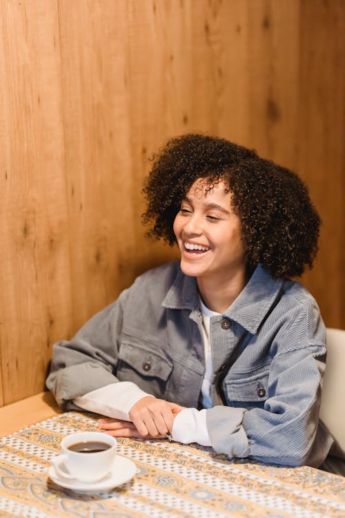 Free Happy young female with curly hair smiling at table with cup of coffee in cafe Stock Photo