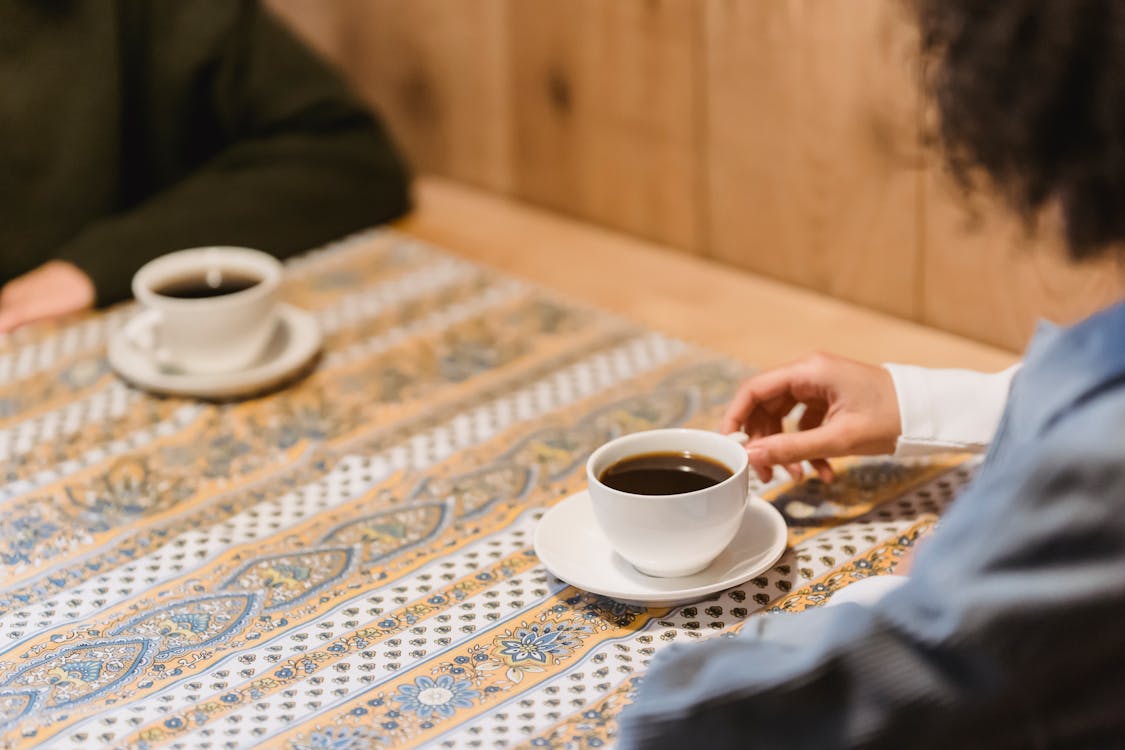 Free From above of crop anonymous women drinking mugs of hot aromatic organic coffee at table with tablecloth with ornaments Stock Photo