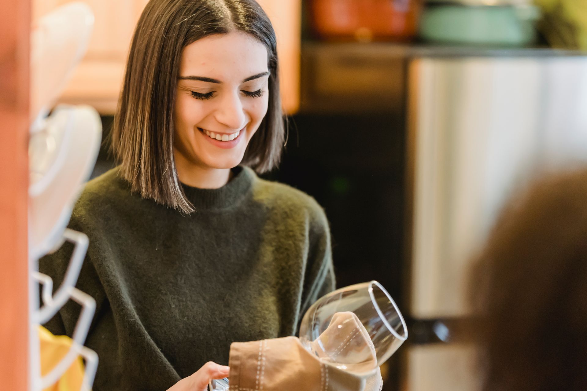 Crop cheerful female smiling and drying clean transparent glass with fabric on blurred background of kitchen
