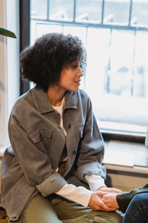 Free Serious young African American woman with dark curly hair in casual clothes holding hand of crop unrecognizable friend sitting near window at home Stock Photo