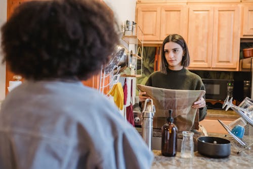 Focused young woman with towel in hand standing in kitchen with anonymous girlfriend