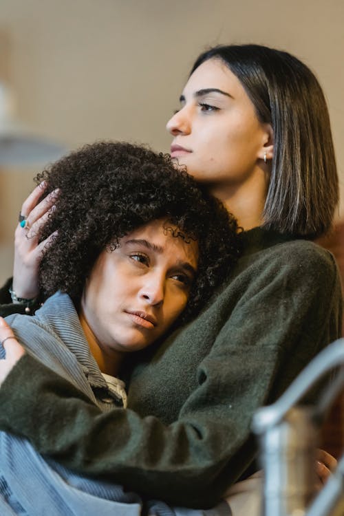 Free Caring female cuddling and stroking head of unhappy African American best friend while standing close and looking away Stock Photo