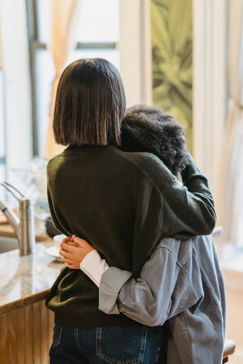 Back view of anonymous woman cuddling and comforting ethnic curly haired girlfriend standing near counter