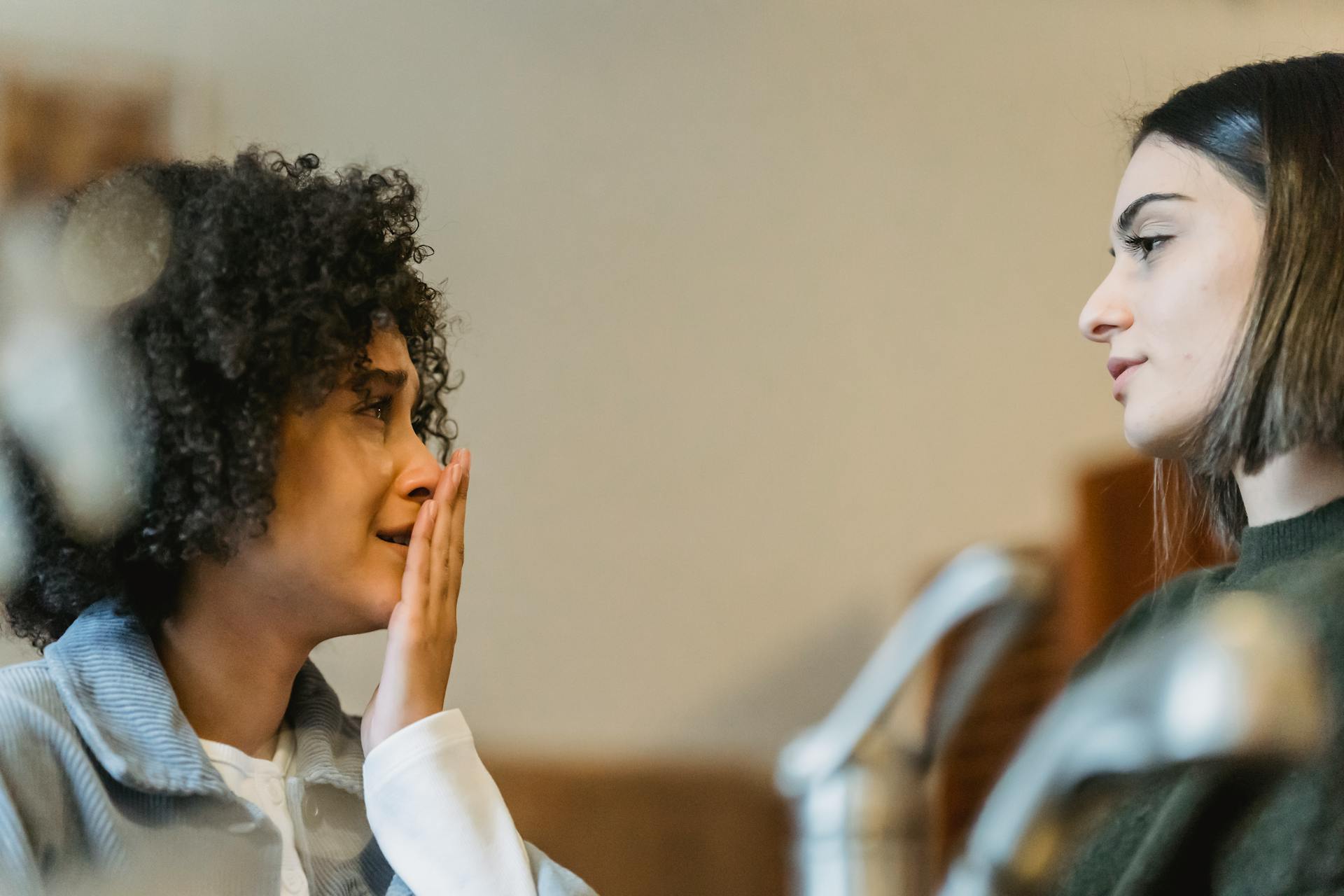 Low angle African American woman covering mouth with hand while crying near supporting girlfriend in kitchen and looking at each other