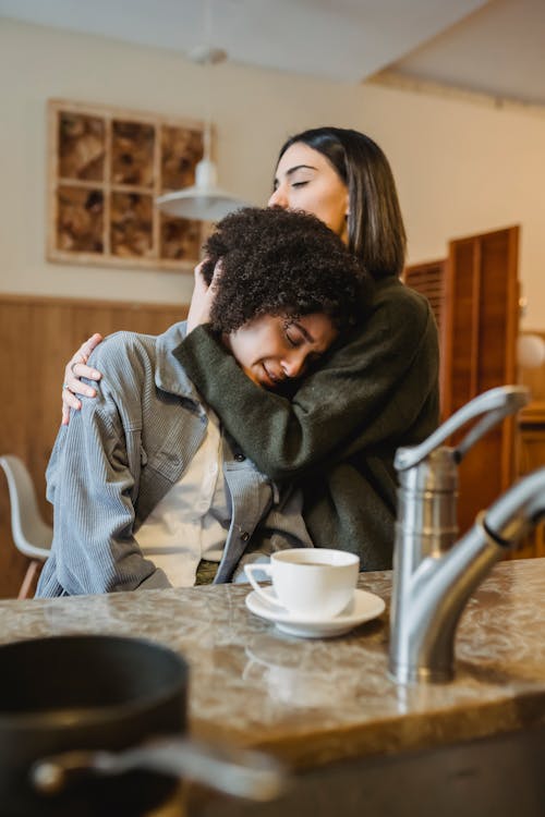 Free Supporting female gently cuddling sad crying African American girlfriend at counter with cup of coffee in kitchen Stock Photo