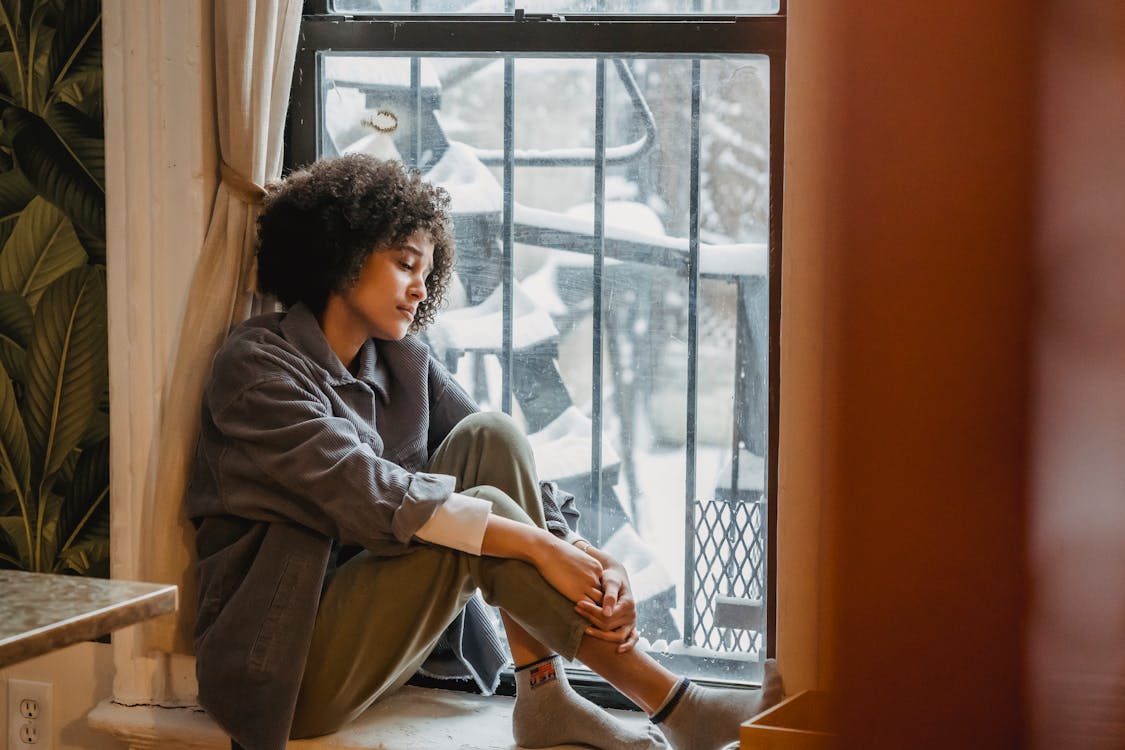 Free Side view full body of upset African American woman embracing knees while sitting in solitude near window and looking down Stock Photo