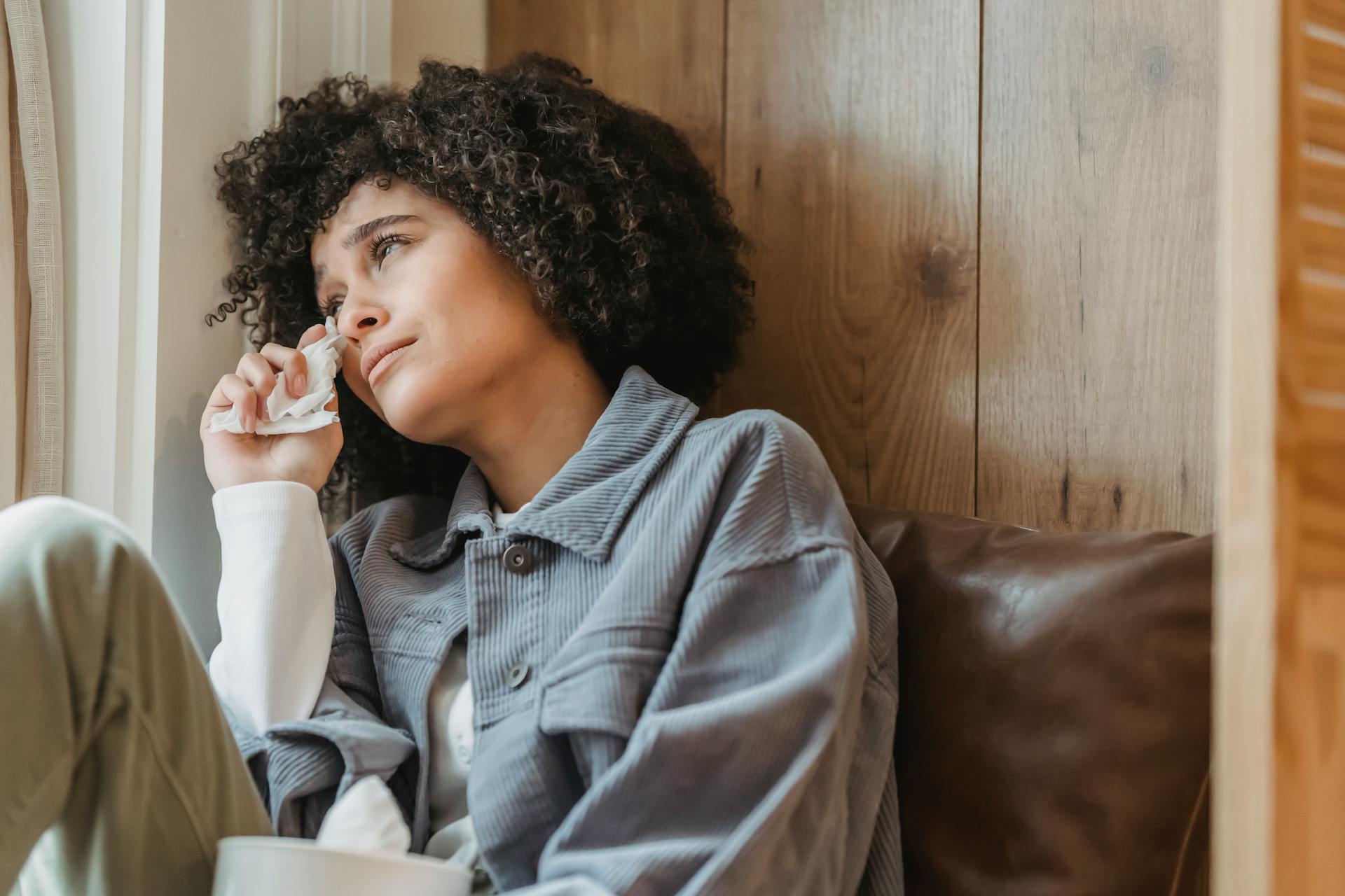 Desperate African American female with curly hair wiping face with tissue while crying from sadness on windowsill and looking away