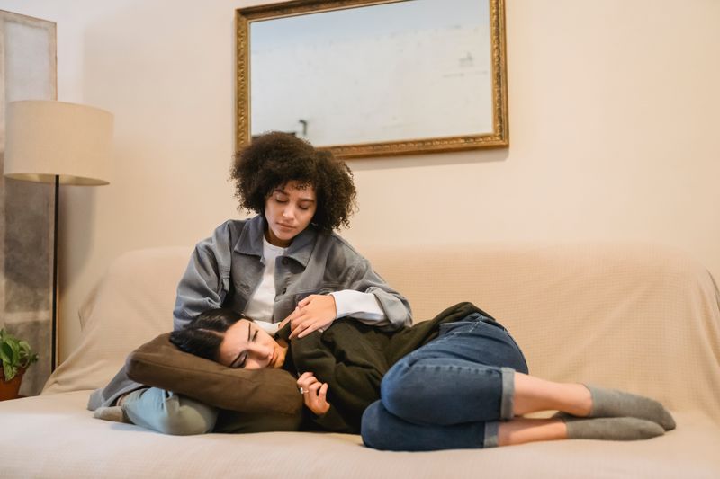 A Black woman sits on a couch comforting a distressed friend who rests across her lap, a portrait of the support and connection that recovery from opioid use disorder depends on.
