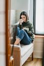 Young melancholic ethnic female millennial in stylish clothes wiping tears with napkins while sitting on couch at home