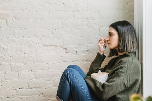 Side view of depressed young ethnic female millennial in warm sweater sitting leaning on wall and wiping tears with tissue at home