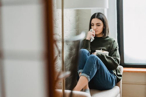 Free Upset young ethnic lady with dark hair in casual clothes sitting on comfortable armchair with tissues box in hand and looking down thoughtfully Stock Photo