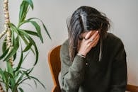 Unrecognizable young upset female millennial with dark hair in stylish sweater covering face with hand while siting on chair at home