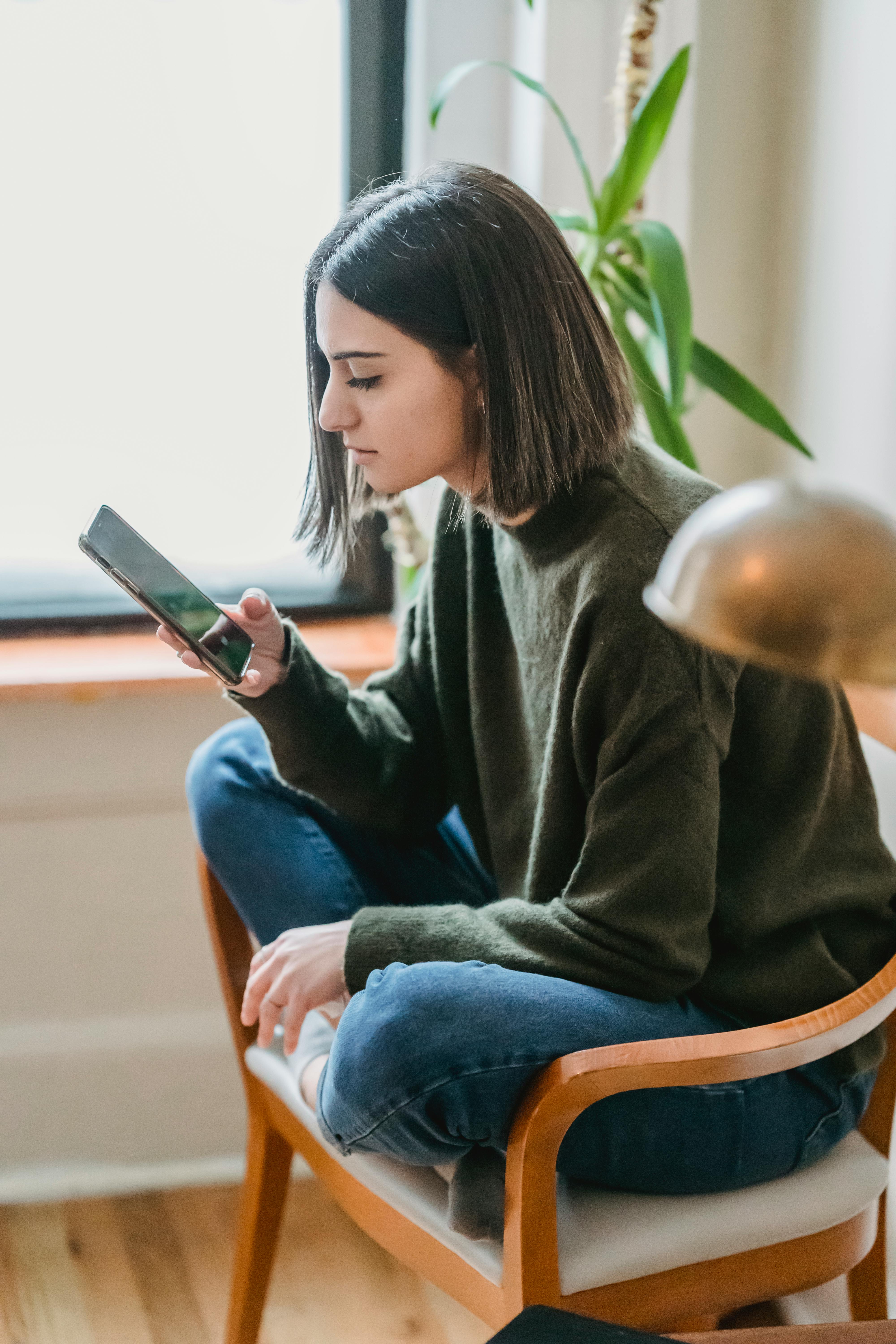 focused young ethnic lady messaging on smartphone sitting on chair