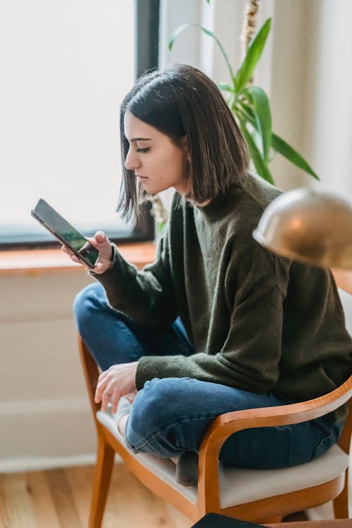 Side view of concentrated young ethnic female millennial sitting on chair with crossed lags and reading message on mobile phone