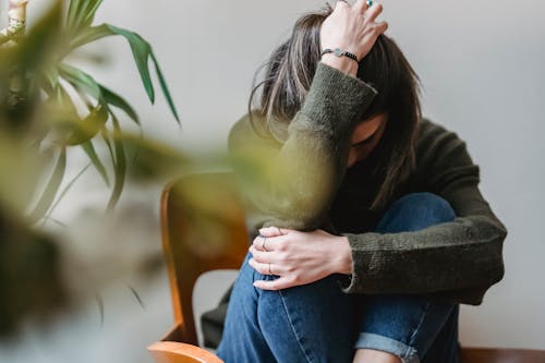 Anonymous young sorrowed female in casual outfit touching dark hair and embracing knees while sitting on chair at home