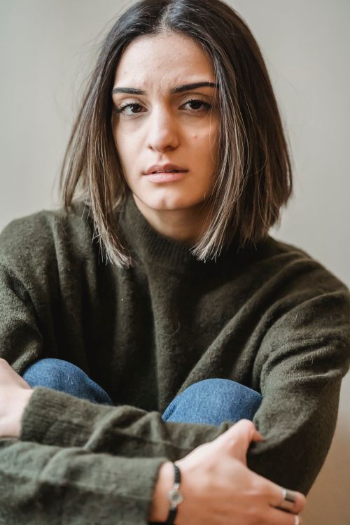 Free Focused young upset ethnic female with stylish haircut in casual outfit embracing knees and looking at camera against white white background Stock Photo