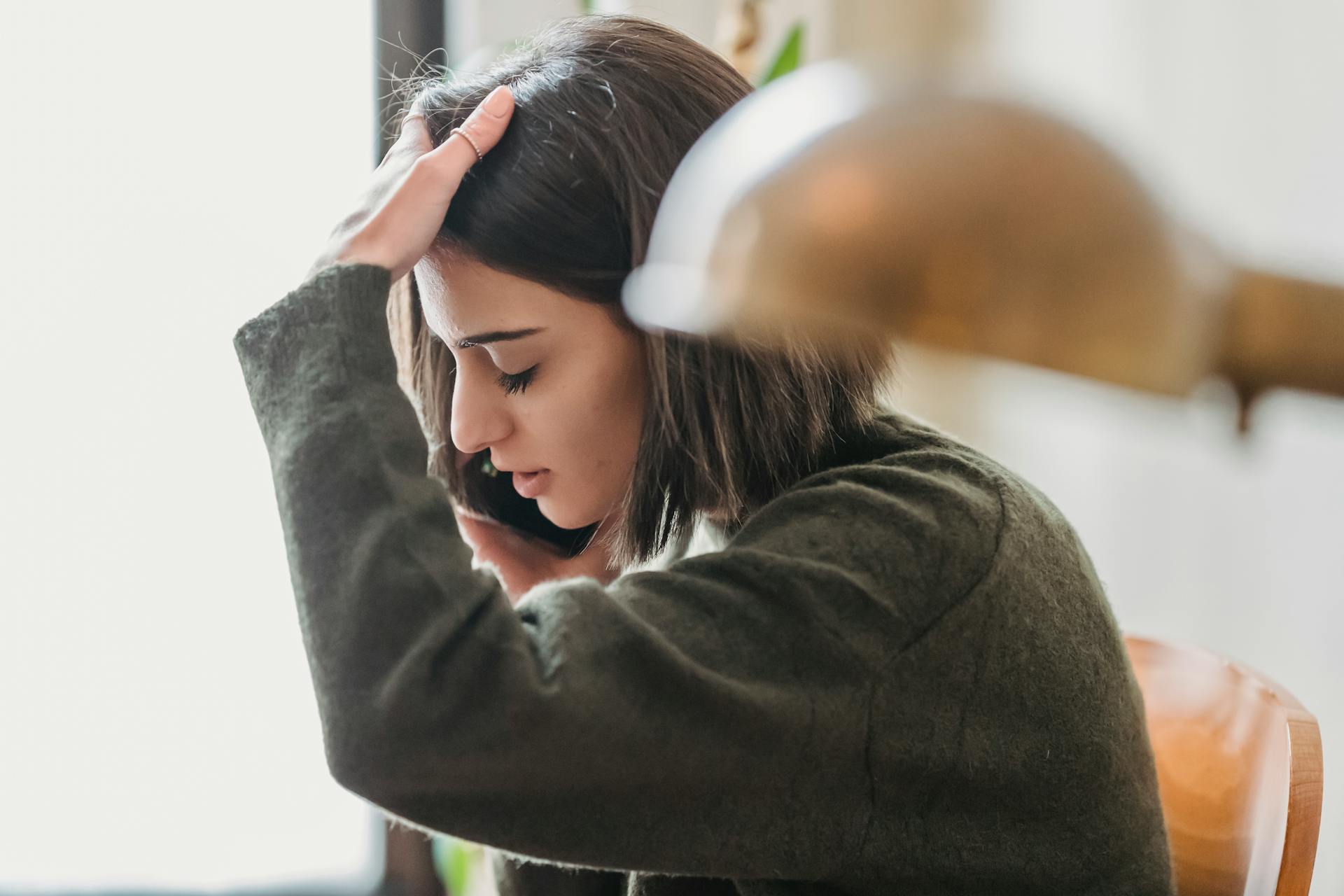 Side view of upset young ethnic female millennial with dark hair grabbing head with closed eyes while having phone conversation sitting on chair at home