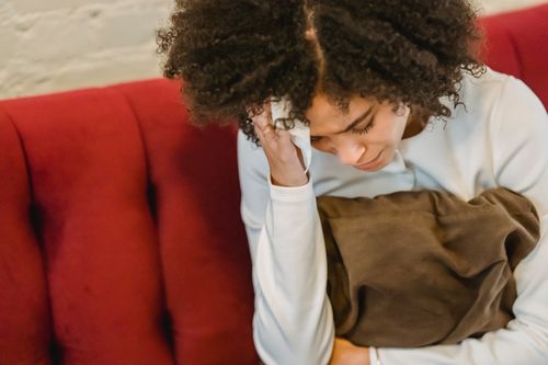 Free From above of crop worried ethnic lady with Afro hair in casual clothes touching head with closed eyes while having headache sitting on sofa at home Stock Photo
