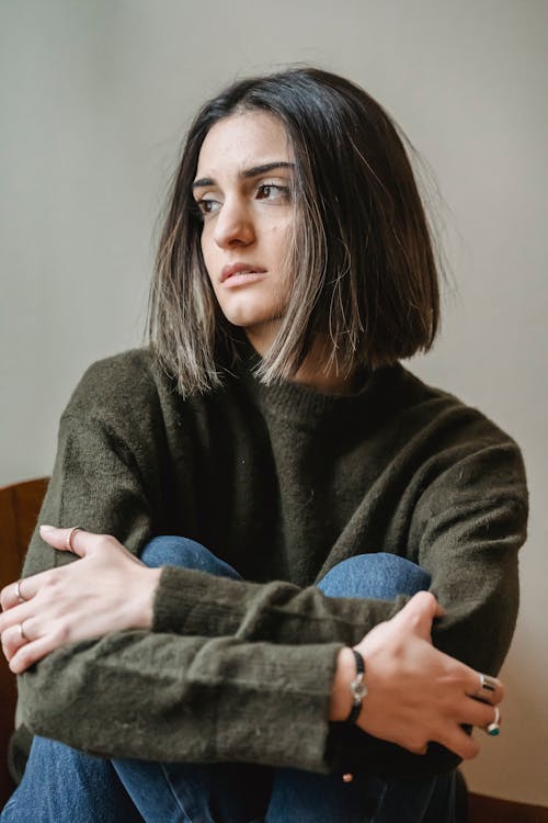 Dreamy young ethnic female millennial with dark hair in casual clothes embracing knees and looking away while sitting alone on chair at home